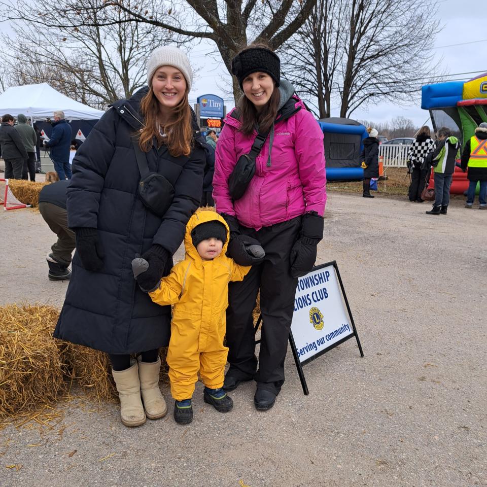 Two female adults with a young child in a bright yellow/orange snow suit smiling for the camera in front of the Tiny Lions Club mini stick arena