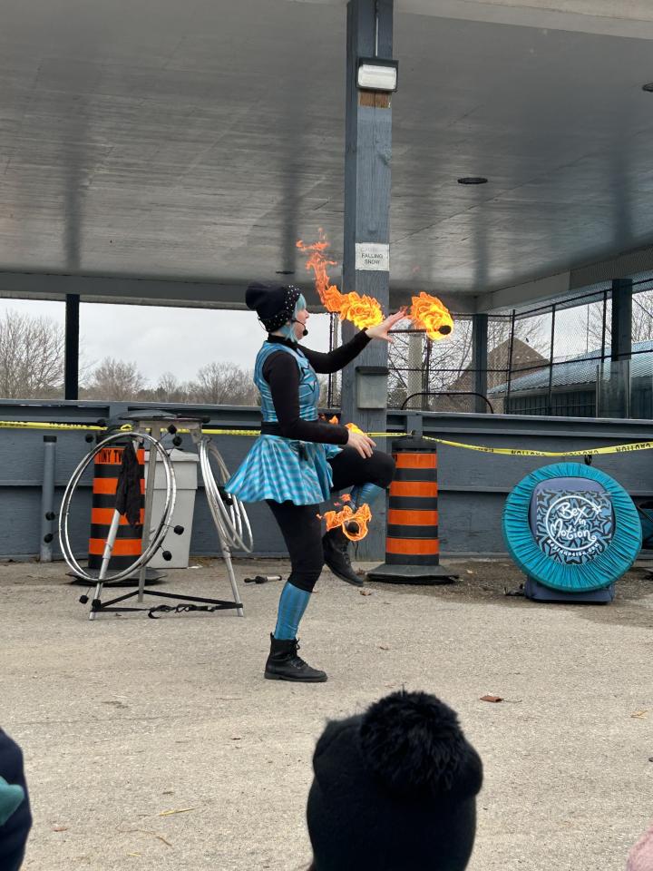 Bex in Motion during their fire performance with a flaming stick in their hand and one resting on their ankle