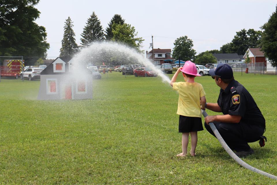 Young child with a firefighter using the fire hose to aim at a mock house fire